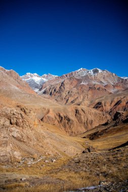 Uzakta engebeli dağlar ve berrak mavi gökyüzü olan uçsuz bucaksız, kahverengi ve altın bir dağ vadisinin dikey görüntüsü.