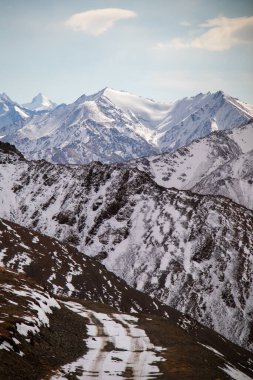 Karla kaplı engebeli bir dağ heybetli karlı bir dağ sırasına doğru uzanan dolambaçlı bir yol bulutlu bir gökyüzünün altında.