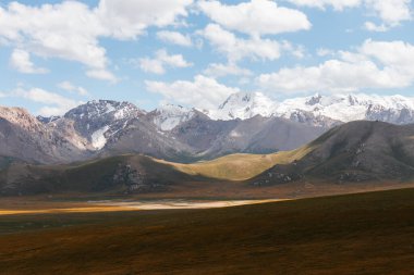 Uçsuz bucaksız, altın ve yeşil bir vadinin panoramik manzarası. Arka planda, bulutlu mavi gökyüzünün altında görkemli kar örtülü dağlar var.