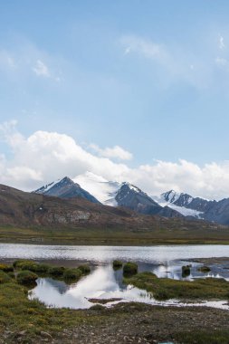 Dağ vadisindeki küçük bir gölün sakin bir görüntüsü. Etrafı karlı dağlarla kaplı yeşil tepelerle çevrili bulutlu bir gökyüzünün altında.