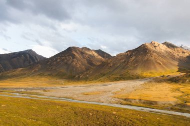 Geniş, altın ve yeşil bir vadinin panoramik manzarası içinden akan bir nehir ve bulutlu bir gökyüzünün altında engebeli dağlar.