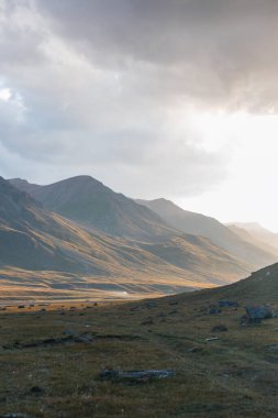 Güneş ışığı dalgalı tepelerin ve engin bir dağ vadisinin üzerinden akıyor. Uzakta bulutlu bir gökyüzünün altındaki heybetli dağların soluk bir manzarası var.