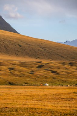 Uçsuz bucaksız, altın bir yamaçta küçük bir yerleşim yeri ve mavi gökyüzünün altında geniş tepeler ve büyük bir dağ var.