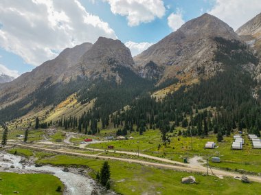 Geleneksel bir yurt kampı manzarası ve yeşil Barskoon vadisinde yuva yapmış küçük binalar, önünde bir nehir ve arkasında yüksek kayalık dağlar.