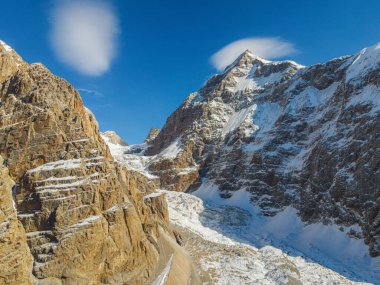 Keskin, karla kaplı bir dağ zirvesinin heybetli manzarası. Zirvesine yakın duran merceksel bir bulut, kayalık bir sırt ve bir buzulun üzerinde süzülüyor.