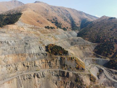 Geniş bir açık çukur madeninin teraslı duvarlarının uçsuz bucaksız ölçeğini ve dokusunu sonbahar ormanı parçalarıyla gösteren yakın çekim bir hava görüntüsü.
