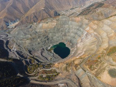 Büyük bir açık çukur madeni, uzak dağ vadisinde görünen küçük bir köyle teraslı yapısı ve merkezi gölü ile araziye hakim.
