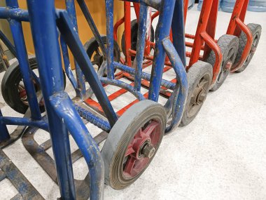 Close-up of stacked blue and red hand trucks with worn wheels in a warehouse setting. Industrial trolleys lined up in storage, showing signs of heavy usage and utility