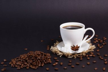 Siyah kahveyle dolu beyaz bir kupa şeker küpleri ve yıldız anasonlu bir fincan üzerinde oturur. Kahve çekirdekleri fincanın etrafına saçılmış ve köşede koyu bir arka planda bir yığın duruyor..
