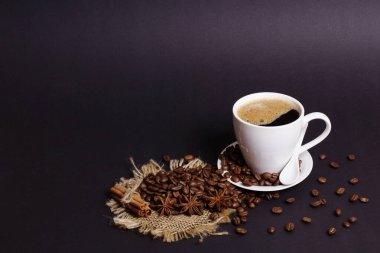 Siyah kahve dolu beyaz bir fincan, kızarmış fasulye, yıldız anason ve tarçın çubuklarının yanında bir fincan üzerinde oturur. Malzemeler bir çuval bezi yüzeyine koyu bir arka plana yerleştiriliyor..