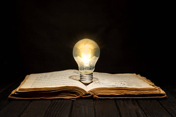 Vintage book with light bulb on wooden table with dark text space.
