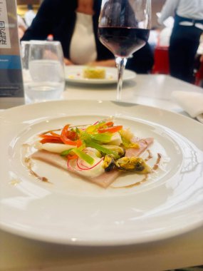 Beyaz bir tabakta sunulmuş gurme deniz ürünleri aperatifi, üzerinde çeşitli kabuklu deniz ürünleri, mürekkep balığı ve renkli dolmalık biber ile pişirilmiş jambon dilimleri var. İnce turp dilimleriyle süslenmiş ve balzamik jölelerle süslenmiş.