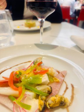 Beyaz bir tabakta sunulmuş gurme deniz ürünleri aperatifi, üzerinde çeşitli kabuklu deniz ürünleri, mürekkep balığı ve renkli dolmalık biber ile pişirilmiş jambon dilimleri var. İnce turp dilimleriyle süslenmiş ve balzamik jölelerle süslenmiş.