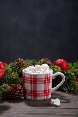 Red mug with hot chocolate on top of which pieces of marshmallows float, and behind are branches of a Christmas tree decorated with red balls, with copy space on wooden background
