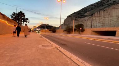 Alicante, İspanya - 25 Nisan 2025: Arkadaşlar karanlık bir gökyüzü altında manzaralı dağlar ve şehir ışıklarıyla çevrili kıyı şeridi boyunca sakin bir akşam yürüyüşünün tadını çıkarıyorlar. Atmosfer huzurlu ve...