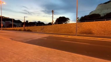 Alicante, İspanya - 25 Nisan 2025: Sersemletici uçurumların yanında, batan güneşten gelen sıcak renklerle yıkanmış sakin bir yolda akşam yürüyüşü başlıyor. Sakin atmosfer yansımayı ve merakı davet eder. Otobüs