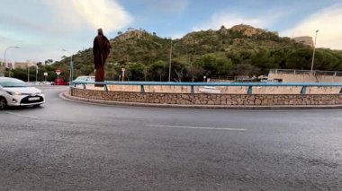 Alicante, İspanya - 25 Nisan 2025: Zarif bir döner kavşakta yemyeşil ve tepelerle çevrili çarpıcı büyük bir heykel sergileniyor. Araçlar geçiyor ve bu kıyıda canlı bir atmosfer yaratıyor.
