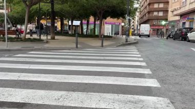 Alicante, İspanya - 25 Nisan 2025: Kalabalık bir kentsel alanda, yayalar yaya geçidinde ilerlerken araçlar durdu. Atmosfer yakınlardaki yerel dükkanlar ve yeşillik kaplamalarıyla dolu.