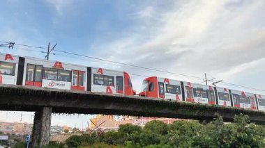 Alicante, İspanya - 25 Nisan 2025: Şehir yeşilliği ve dramatik bulutlu gökyüzü arasında yükseltilmiş bir tren yolunda sorunsuzca ilerliyor. Bu sahnede modern ulaşım sergileniyor.