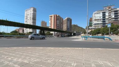 Trafik kıyı şehrinde hareketli bir kavşaktan geçiyor, modern binalar sergileniyor ve açık mavi gökyüzüne karşı hareketli bir ufuk çizgisi.