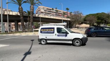 Alicante, İspanya - 29 Mayıs 2025: Arabalar mavi gökyüzü altında işlek bir caddede yol alırlar. Palmiye kuleleri yukarıda şehir yapıları yakınlarda gezinirken, şehir yaşamının davetkar atmosferini yaratıyor..