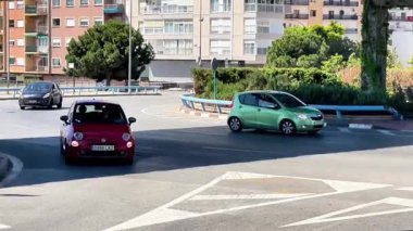 Açık gökyüzünün altında hareketli bir şehir ortamında iki araba dolambaçlı bir yoldan gidiyor. Yeşil araç başka bir şeritten yaklaşırken kırmızı araba çıkışa doğru ilerliyor..