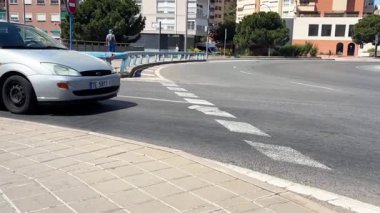 Alicante, İspanya - 29 Mayıs 2025: Bir kişi aydınlık bir günde kaldırım boyunca yürürken bir araba döner kavşakta pürüzsüz döner. Kentsel çevrede modern binalar ve tabelalar yer alıyor.