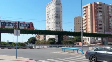 Güneşli bir günde, hareketli bir şehir manzarasında, yüksek binalar arasındaki boşluğu dolduran, yükseltilmiş bir ray boyunca bir tren sorunsuzca hareket eder..