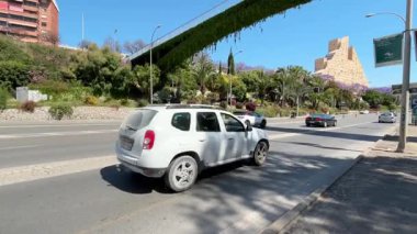 Güneşli bir günde, araçlar yemyeşil ve renkli çiçeklerle bezenmiş canlı bir sokaktan geçer. Yerin cazibesini ve canlı atmosferini sergilerler..