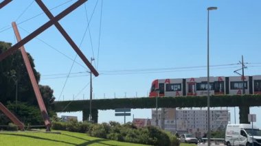 Tramvay, parlak mavi gökyüzünün altında modern mimarinin yer aldığı yeşil bir çizgi üzerinde zarifçe hareket ediyor. Renkli bir şehir heykeli canlı şehir atmosferini artırır.