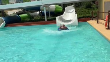 Alicante, İspanya - 13 Haziran 2025: Genç çocuk neşeyle havuza kayıyor, açık mavi su ve güneşli atmosferle çevrili.