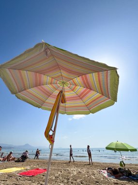 Alicante, İspanya - 24 Haziran 2025: Renkli çizgili plaj şemsiyesi kumlu plajda gölge sunarken güneşlenenler dinleniyor ve sıcak yaz gününün tadını çıkarıyorlar