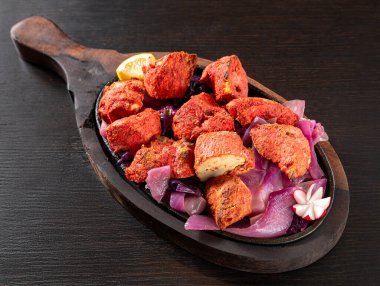 Kırmızı Murg Tikka 'nın yakın çekimi, yoğurt, otlar ve baharatlarla marine edilmiş geleneksel Hint tavuk parçaları, soğanla birlikte cızırdayan bir tabakta servis edilir, koyu bir ahşap arka planda limon dilimi ile izole edilir.