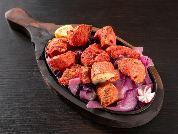 Kırmızı Murg Tikka 'nın yakın çekimi, yoğurt, otlar ve baharatlarla marine edilmiş geleneksel Hint tavuk parçaları, soğanla birlikte cızırdayan bir tabakta servis edilir, koyu bir ahşap arka planda limon dilimi ile izole edilir.