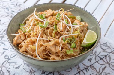 Pirinç erişteli, sebzeli, tavuklu ve demirhindi soslu Pad Thai tabağı dekore edilmiş masa örtüsü arka planında yeşil bir seramik kasede servis ediliyor. Parlak fotoğraf, Tayland mutfağı teması için ideal.