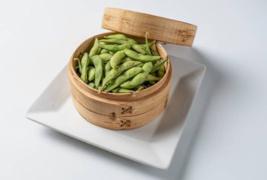 Geleneksel Çin bambu buharlı buharlı bir tenceredeki taze edamame, beyaz minimalist arka planda beyaz bir tabağa yerleştirilir. Temiz ve doğal bir estetik ile sağlıklı Asya mutfağını sergilemek için mükemmel..