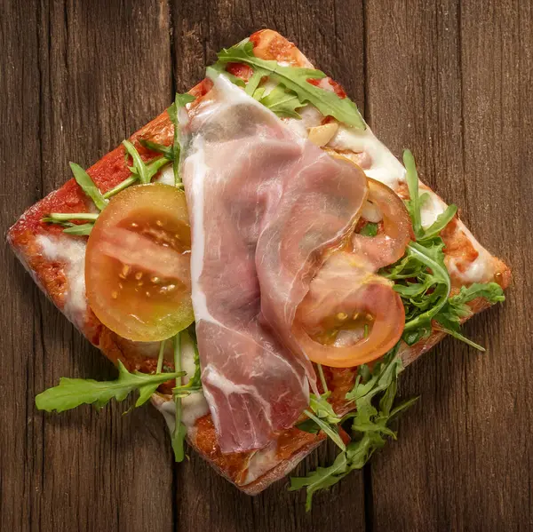Domatesli İtalyan pizza dilimi, çiğ jambon, mozzarella ve roket kırsal ahşap masada. Geleneksel tarz, parlak renkler, basit kompozisyon, iştah açıcı yemek.