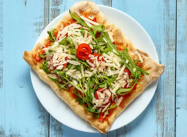 Beyaz tabakta domates, roket ve parmesan peynirli dilimlenmiş İtalyan pizzası. Eski tarz, açık mavi ahşap bir masada. Kırsal stil, parlak renkler, basit kompozisyon 