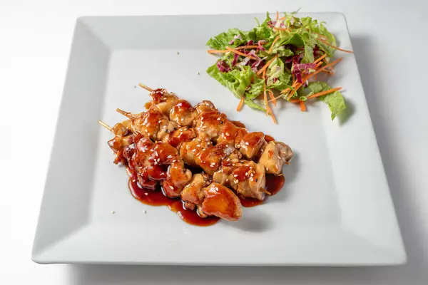 Geleneksel tavuk yakitori, Japon soslu ızgara tavuk şişi, beyaz arka planda beyaz bir tabakta salata ve sosla servis ediliyor. Temiz, canlı estetik bir Asya yemeği..