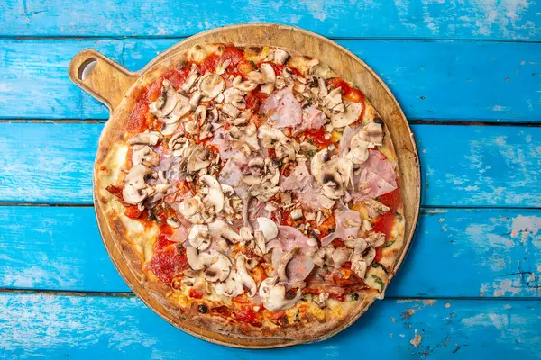 Domatesli, mozarellalı, jambonlu ve mantarlı ekstra büyük İtalyan pizzası. Mavi, pejmürde bir arka planda izole edilmiş. Kırsal, canlı bir fotoğraf, İtalyan mutfağı ve pizza temaları için mükemmel.