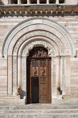 İtalya, Umbria 'daki Foligno Katedrali' nin ortaçağ oymalı yan kapısı. Pembe mermer bantlı Romanesk Gotik kemer, karmaşık oymalar ve güneşli ahşap kapı Foligno, İtalya, 12 Temmuz 2025