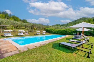 Güneşli bir yaz gününde güneşli bir yaz gününde güneşli yataklar, güneş şemsiyeleri ve zeytin ağaçlarıyla dolu, yeşil tepelerle çevrili kırsal bir villada yüzme havuzu..