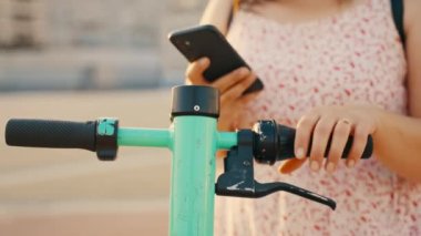Sırt çantalı genç bir kadın internet üzerinden kiralık yeşil bir scooter 'ı çalıştırmak için akıllı telefonunu kullanıyor. Yüksek kalite 4k görüntü