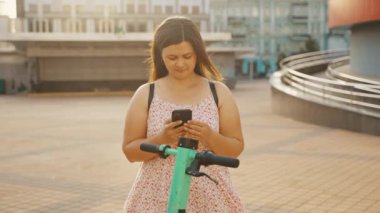 Elbiseli genç bir kız, uygulamadan açık yeşil elektrikli scooter kiralamak için akıllı telefonunu kullanıyor. Özetle elektrikli scooter 'a binmek. Yüksek kalite 4k görüntü