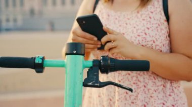 Sırt çantalı genç bir kadın internet üzerinden kiralık yeşil bir scooter 'ı çalıştırmak için akıllı telefonunu kullanıyor. Yüksek kalite 4k görüntü