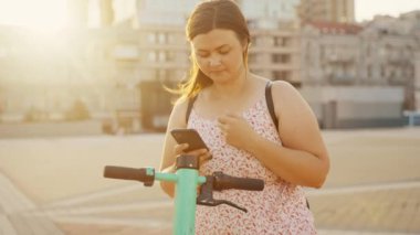 Sırt çantalı genç bir kadın internet üzerinden kiralık yeşil bir scooter 'ı çalıştırmak için akıllı telefonunu kullanıyor. Yüksek kalite 4k görüntü