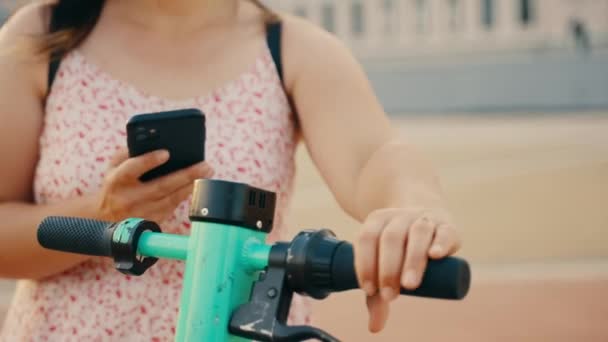 バックパック付きのドレスを着た若い女性は 路上でスマートフォンを使用してインターネット経由でレンタルした通りの緑の電動スクーターをアクティブにします 高品質4K映像 — ストック動画