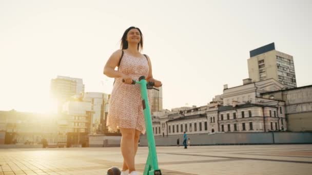 街の風景を通して晴れた日に電動スクーターに乗って陽気な女の子のスローモーション 電動スクーターに乗って 4K判の高画質撮影 — ストック動画