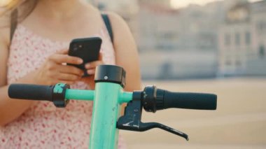 Sırt çantalı genç bir kadın internet üzerinden kiralık yeşil bir scooter 'ı çalıştırmak için akıllı telefonunu kullanıyor. Yüksek kalite 4k görüntü