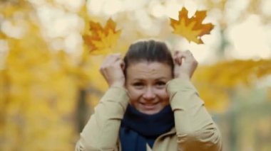 Genç bir kız ellerinin önünde yaprakları tutuyor, onları hareket ettiriyor ve komik suratlar yapıyor. Temiz havada yürü. Autumn Park 'ta. Yüksek kalite 4k görüntü